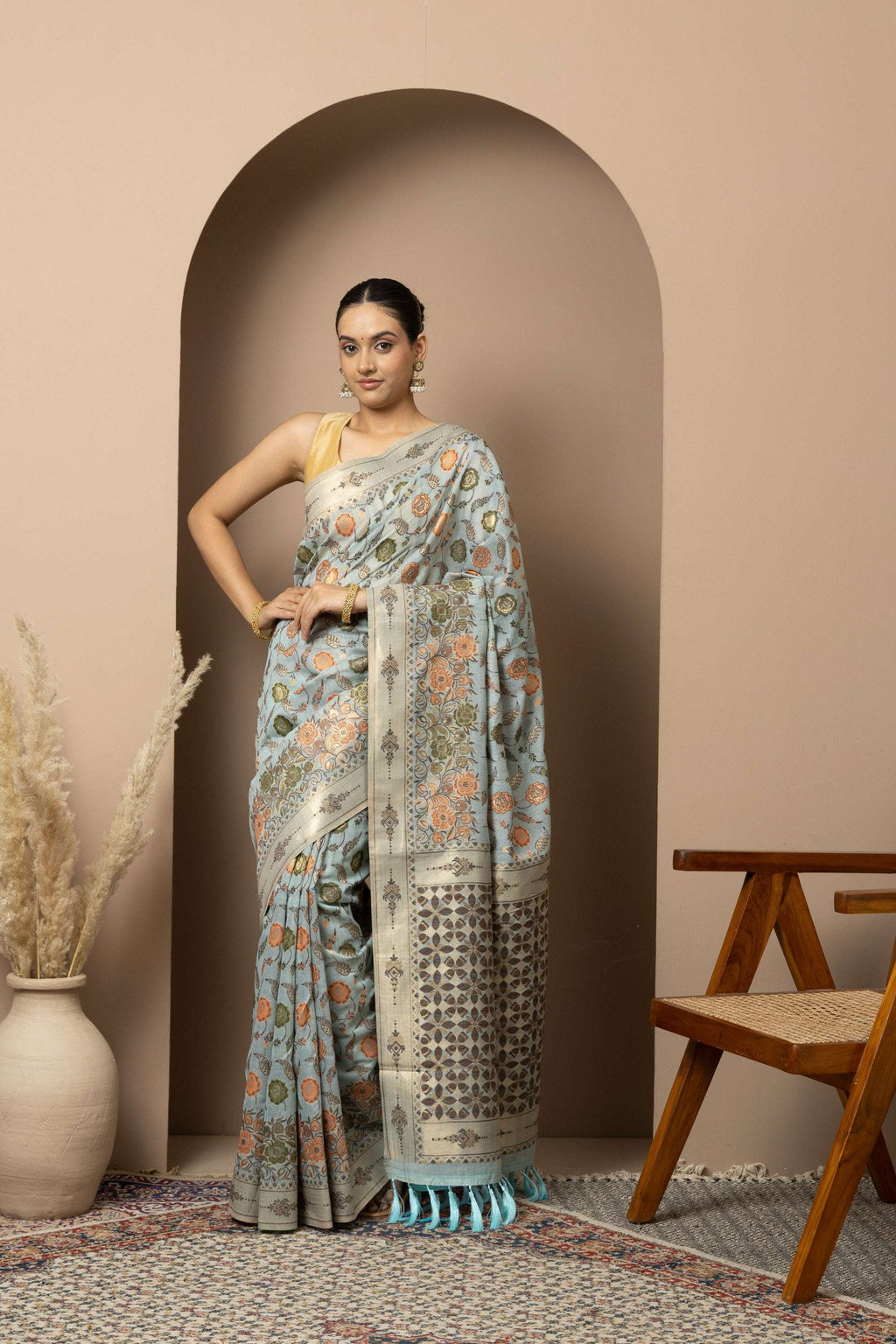 Woman wearing a floral saree standing in front of an archway with a wooden chair and vase in the background.